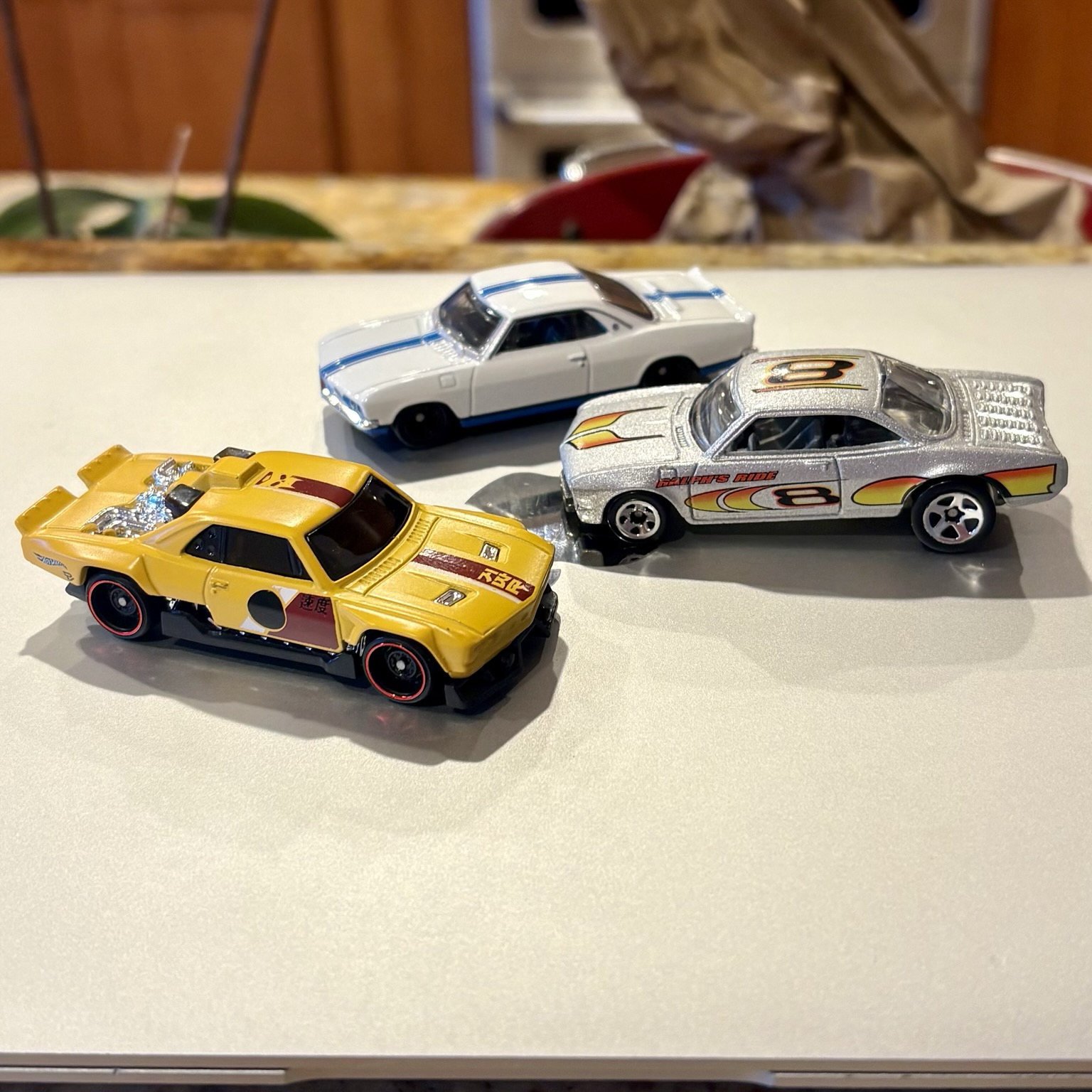A trio of Hot Wheels cars, all customized late-model Chevrolet Corvair coupes, sit atop a closed silver laptop computer. Clockwise from left: the yellow "Vairstrike," with an exposed chromed mid-mounted engine, released in 2026; a white 1966 Yenko Stinger with blue racing stripes, released in 2022; and a silver "Vairy 8," with a louvered rear deck and a mid-mounted engine molded into the interior, released in 2002.