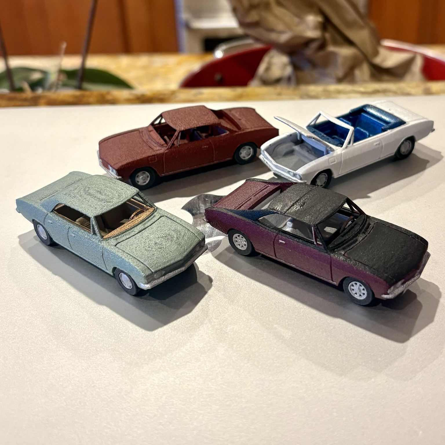 Four 3D printed 1:64-scale Chevrolet Corvair model cars sit atop a closed silver laptop computer. Clockwise from left: a Willow Green 1965 Monza hardtop sedan; an Aztec Bronze 1966 Corsa hardtop coupe; a white 1967 Monza convertible with its front trunklid open; a two-tone burgundy and black 1965 Fitch Sprint hardtop coupe.