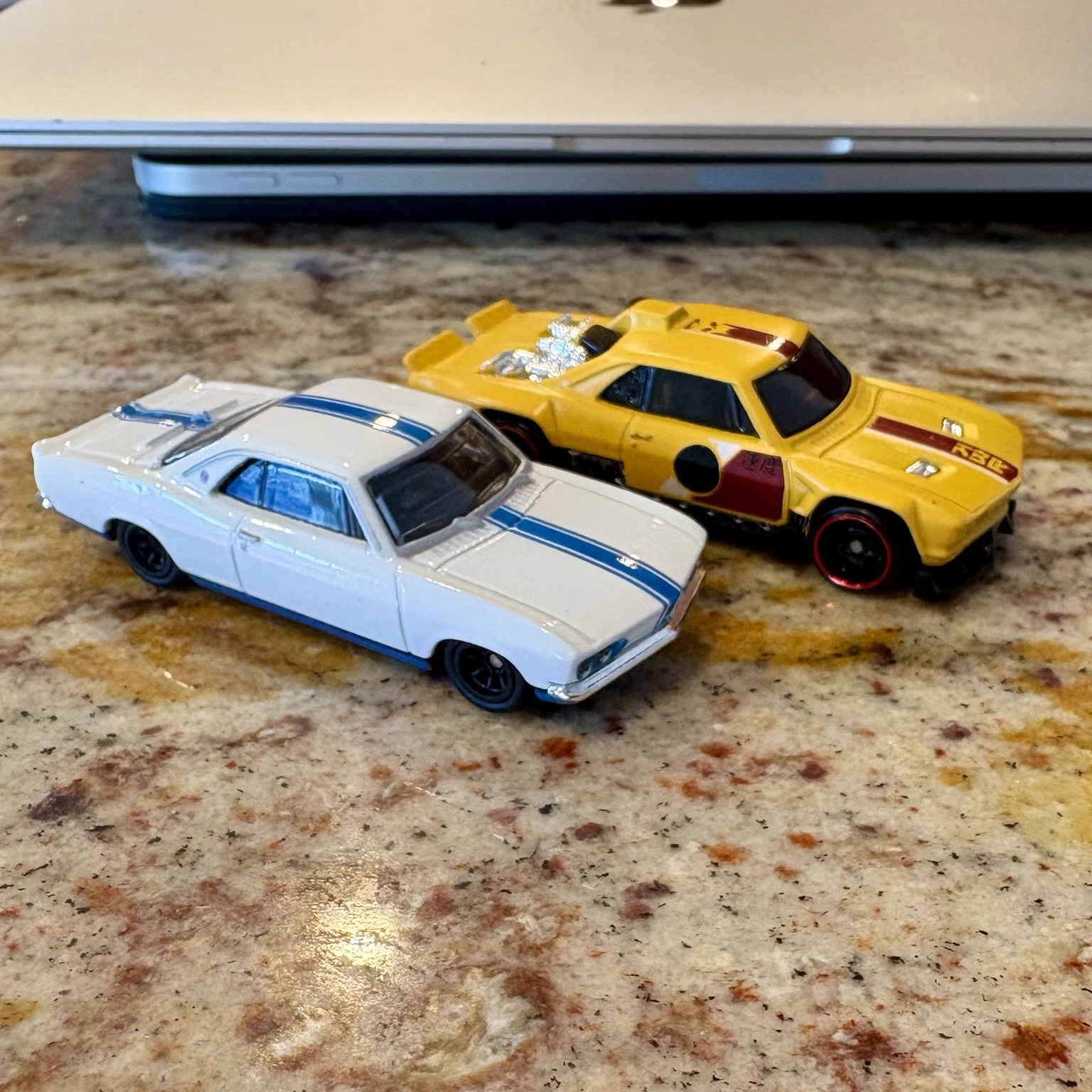 Two Hot Wheels toy cars sit side by side on a brown granite countertop. On the left, a white 1966 Chevrolet Corvair Yenko Stinger coupe with blue racing stripes; on the right, the "Vairstrike," a wildly customized yellow Corvair coupe with a chromed "engine" sticking out where the rear window would normally be.