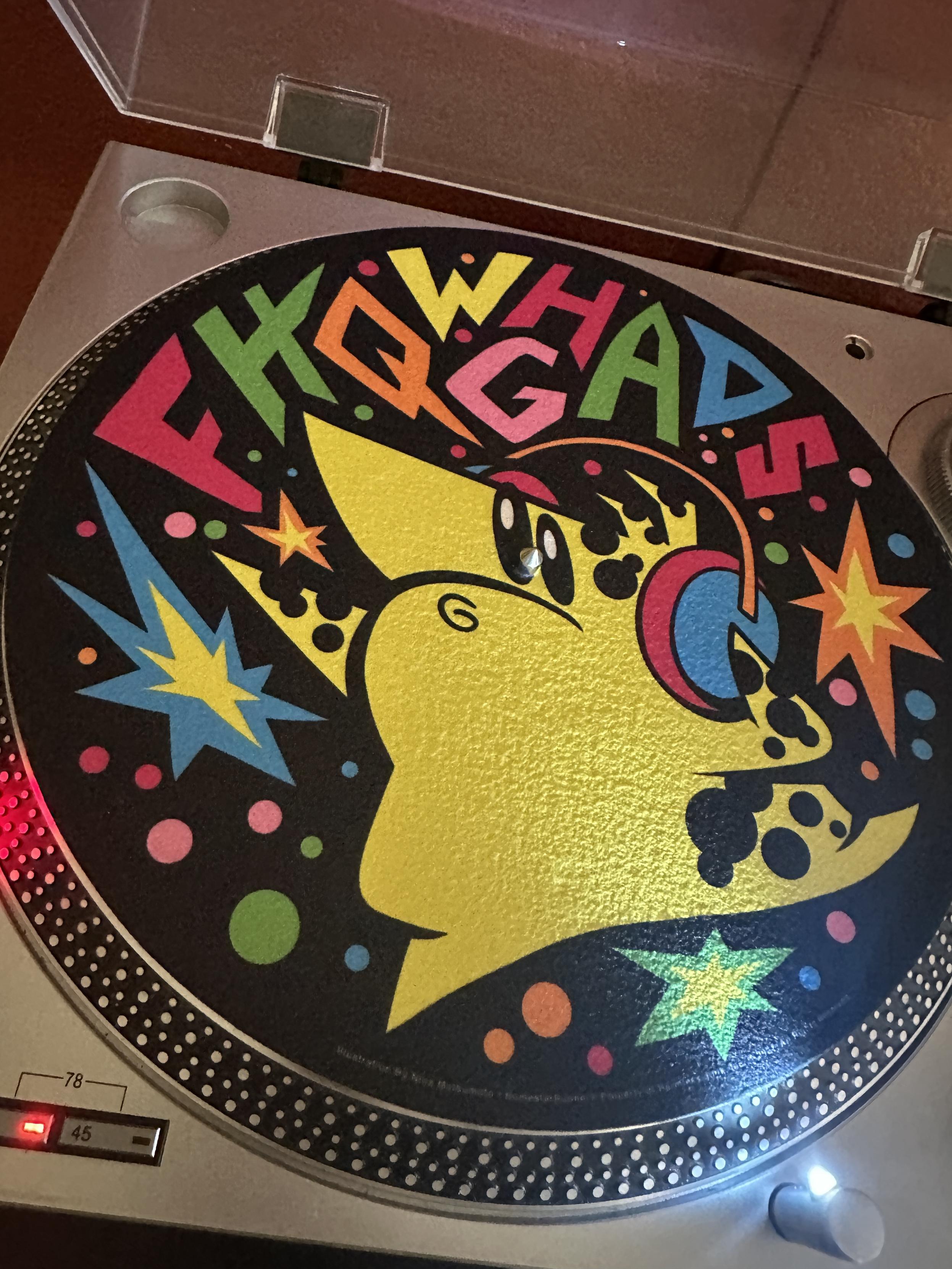 A turntable viewed from the top.  Visible is a bright yellow cartoon character with black spots and headphones on a black background with colorful sparkles and the word "FHQWGHADS"
