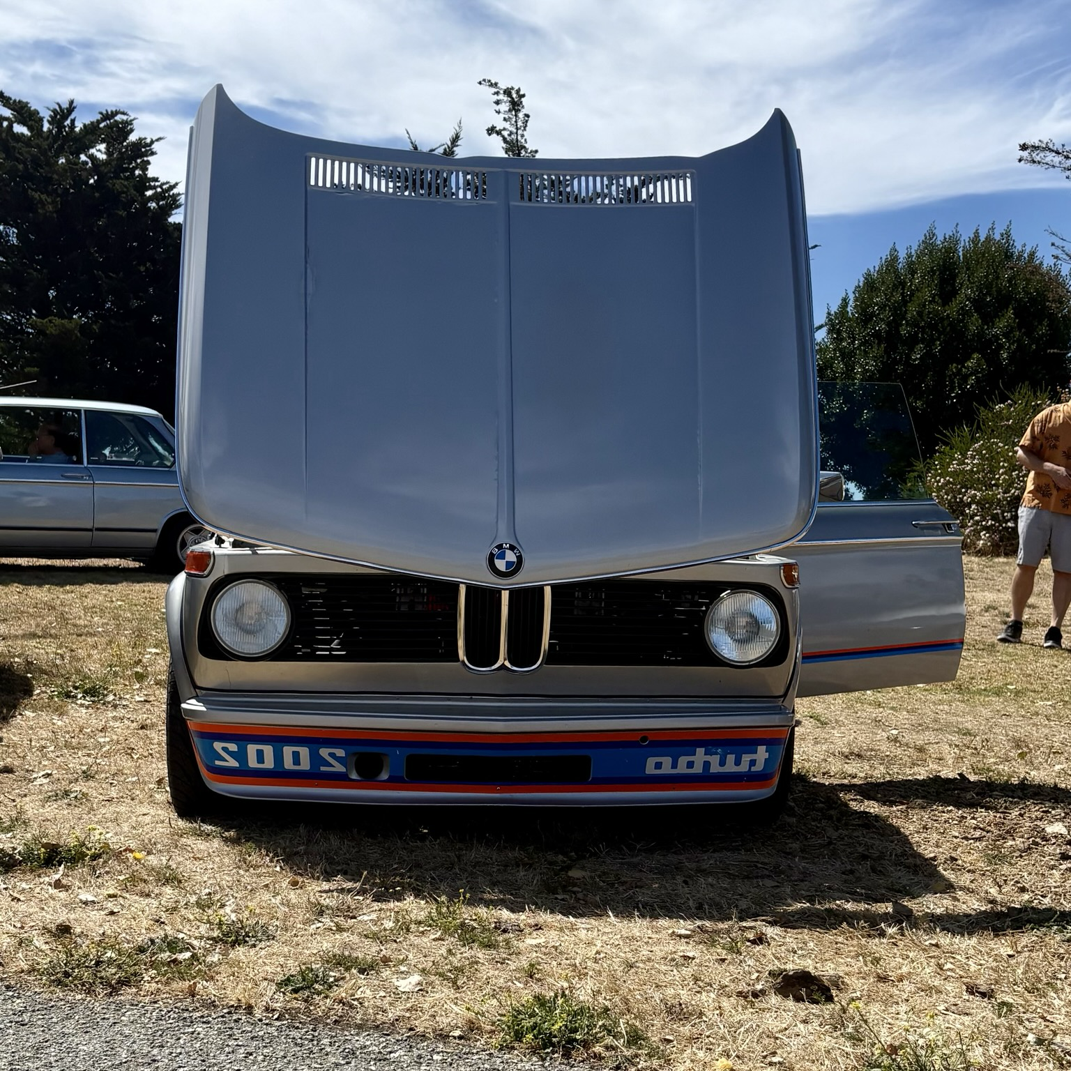 A front view of a silver 1975 BMW 2002 Turbo with its hood and driver door open.