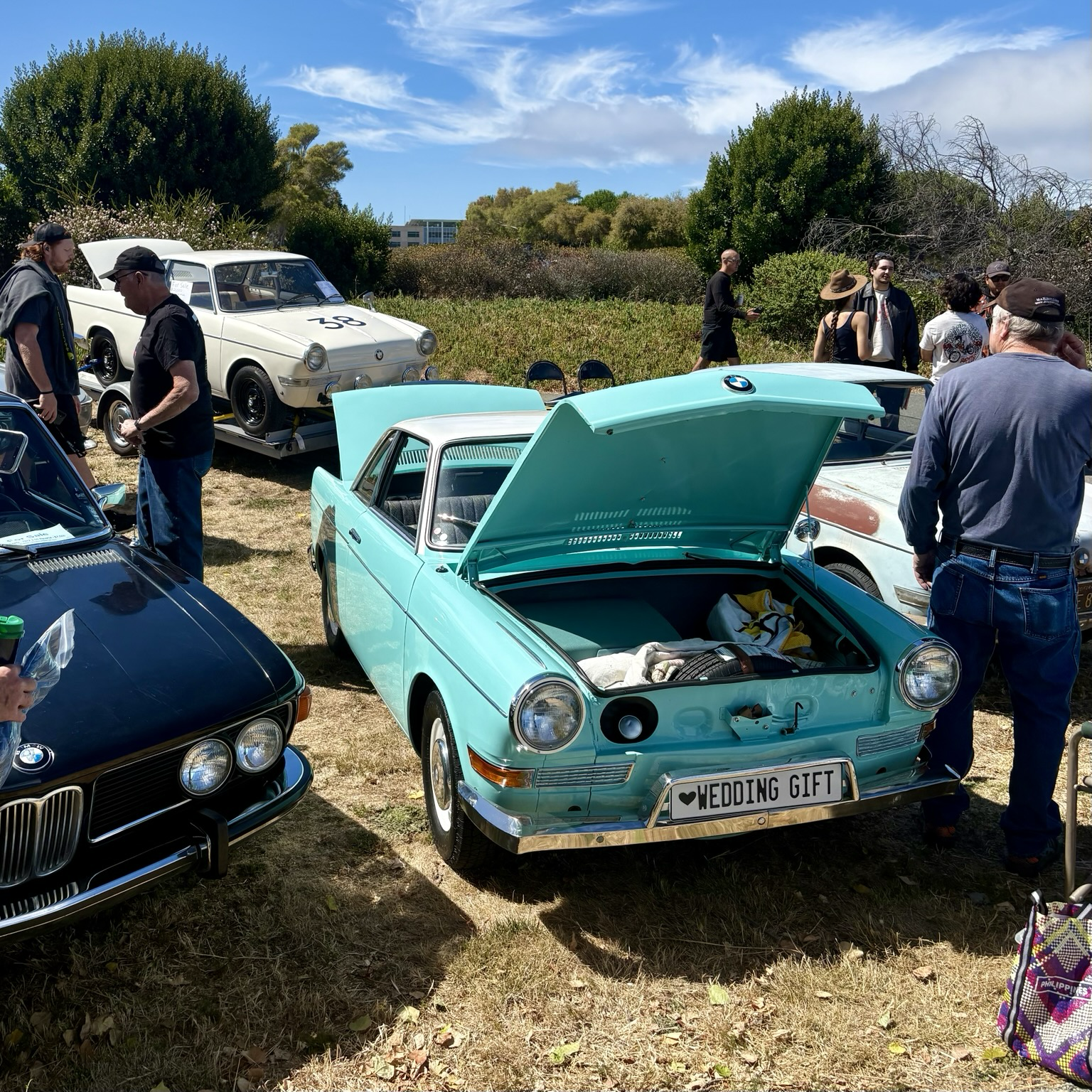 A two-tone aqua and white early 1960s BMW 700 sport coupe with its front and rear deck lids open. A second, white 700, sitting on a trailer, is visible in the background.